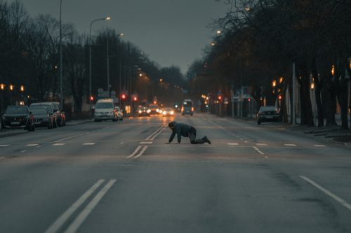Cilvēks rāpo pa ielu krēslā, automašīnas fonā.