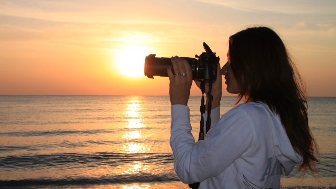 Sieviete fotografē saulrietu pie jūras ar kameru.